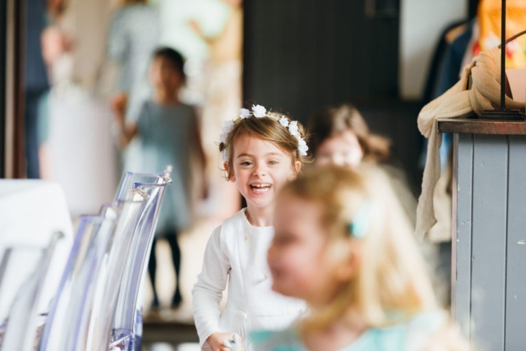 Une jeune fille avec une couronne de fleurs et une robe blanche sourit en marchant dans un intérieur lors d'un baptême mémorable à Bordeaux. Elle est suivie par d'autres enfants, flous en arrière-plan. Le décor semble festif, avec des lumières et des décorations visibles. Elle est près de chaises transparentes et d'une structure en bois, avec une atmosphère chaleureuse qui les entoure.