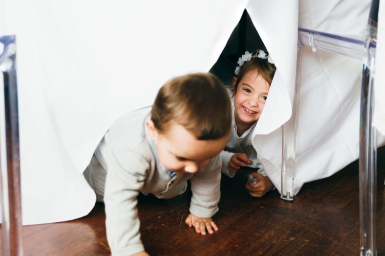 Deux enfants jouent sous une nappe blanche drapée sur une table. L'enfant au premier plan, concentré sur sa marche à quatre pattes, porte une chemise claire. Derrière eux, un enfant souriant avec un bandeau fleuri sort de sous la nappe. Le décor semble se dérouler à l'intérieur sur un plancher en bois, rappelant un baptême mémorable à Bordeaux.