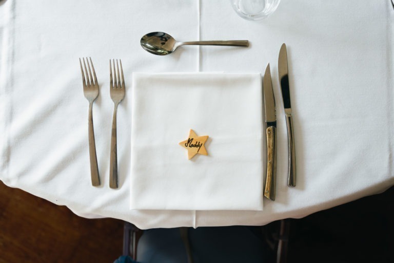 Une table à manger est dressée avec une nappe blanche. On y trouve deux fourchettes à gauche, une cuillère et un couteau à droite, et une serviette blanche pliée au centre. Un marque-place en bois en forme d'étoile avec l'inscription « Papa » dessus orne la serviette. Un verre est partiellement visible dans le coin supérieur droit, parfait pour un baptême mémorable à Bordeaux.