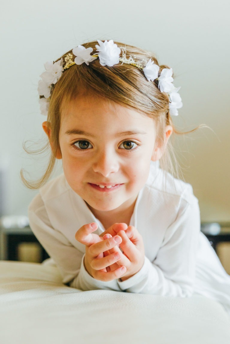 Un jeune enfant aux cheveux bruns ornés d'un bandeau à fleurs blanc sourit chaleureusement à la caméra. Il porte une tenue blanche et joint les mains, marquant un baptême mémorable à Bordeaux. Le fond neutre garantit que l'enfant reste le point central de l'image.