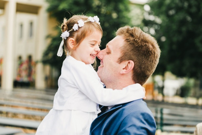 Un homme en costume bleu et chemise blanche tient amoureusement une jeune fille vêtue d'une tenue blanche avec un bandeau fleuri. Ils sont nez à nez, tous deux souriant joyeusement, capturant l'essence d'un baptême mémorable à Bordeaux. L'arrière-plan est flou, montrant des bancs et des arbres verts, suggérant un décor extérieur.