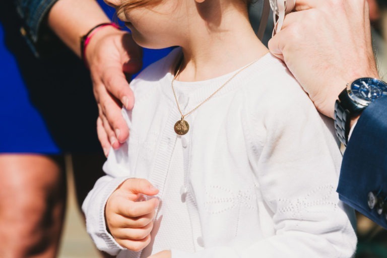 Un jeune enfant, vêtu d'un gilet blanc et d'un collier en or avec un pendentif rond, est délicatement tenu par deux adultes. L'un, à gauche, touche l'épaule de l'enfant tandis que l'autre, à droite, ajuste le collier avec une main portant une montre-bracelet. C'était un véritable baptême mémorable à Bordeaux.