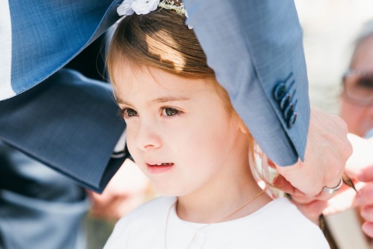 Un jeune enfant aux cheveux châtain clair et coiffé d'un bandeau fleuri se tient immobile tandis qu'un adulte en costume bleu l'aide à mettre un collier. L'enfant a l'air concentré et calme. Une lumière naturelle vive illumine la scène depuis la gauche, mettant en valeur le visage de l'enfant et les détails du costume lors de ce baptême mémorable à Bordeaux.