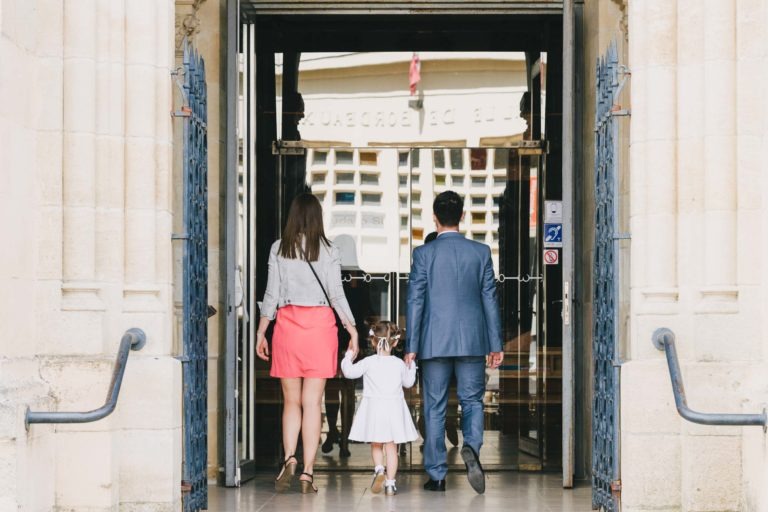 Un homme en costume bleu, une femme en veste grise et jupe rose et une jeune fille en robe blanche et petit sac à dos marchent main dans la main dans l'entrée du bâtiment. L'entrée, encadrée par des murs en pierre beige, évoque une élégance digne d'un château, capturant le charme de leur baptême mémorable à Bordeaux.