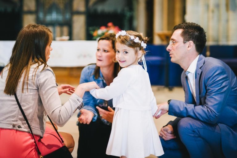 Une jeune fille vêtue d'une robe blanche et d'une couronne de fleurs se tient debout, souriante, tenant la main de trois adultes lors d'un baptême mémorable à Bordeaux. Deux femmes sont agenouillées à sa gauche et un homme en costume bleu est agenouillé à sa droite. Ils sont à l'intérieur d'une église, avec un autel blanc et un bouquet de fleurs visibles en arrière-plan.