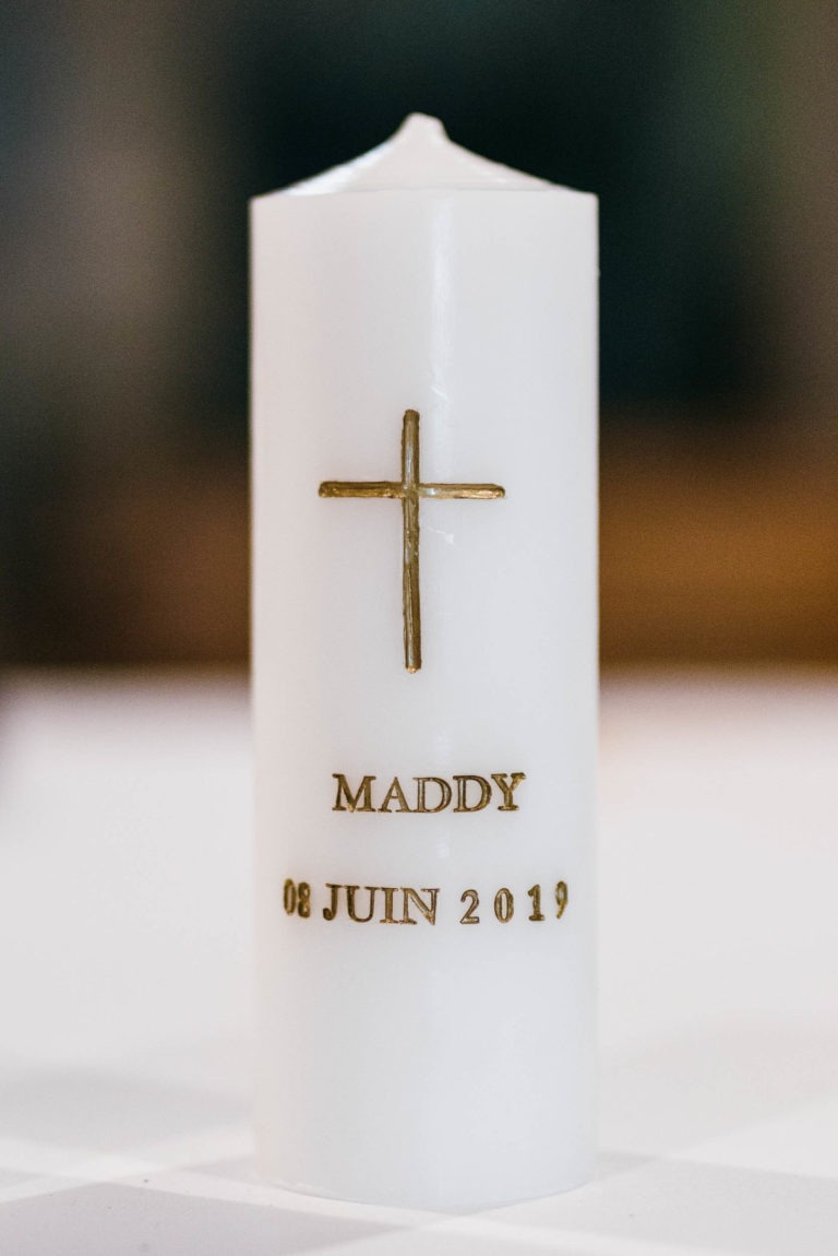 Bougie cylindrique blanche avec une croix en or gravée sur sa face avant, commémorant un baptême mémorable à Bordeaux. Sous la croix, le nom " MADDY " et la date " 08 JUIN 2019 " sont inscrits en or. L'arrière-plan est flou, mettant en valeur la bougie comme point central de l'image.