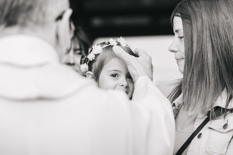 Un prêtre en robe blanche pose délicatement sa main sur la tête d'un jeune enfant, coiffé d'une couronne de fleurs. L'enfant lève les yeux avec une expression calme. Une femme aux cheveux mi-longs, peut-être la mère, se tient à proximité, observant l'interaction avec un comportement chaleureux. Ce baptême mémorable à Bordeaux respire la sérénité et l'intimité.