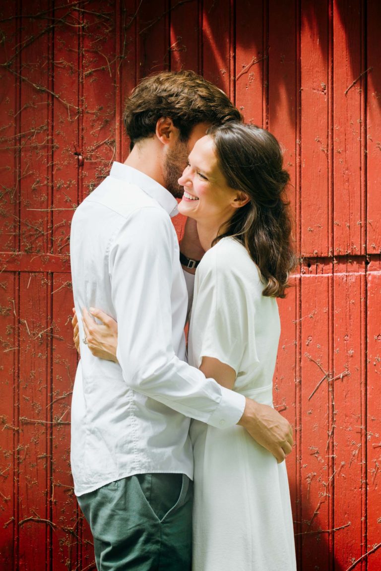 Un couple s'embrasse affectueusement devant un mur en bois rouge dans cette belle séance photo de couple. L'homme, vêtu d'une chemise blanche et d'un pantalon vert, a le dos partiellement tourné vers la caméra. La femme, vêtue d'une robe blanche, sourit et le regarde, son visage illuminé par la lumière du soleil tandis que des vignes grimpent sur le mur.