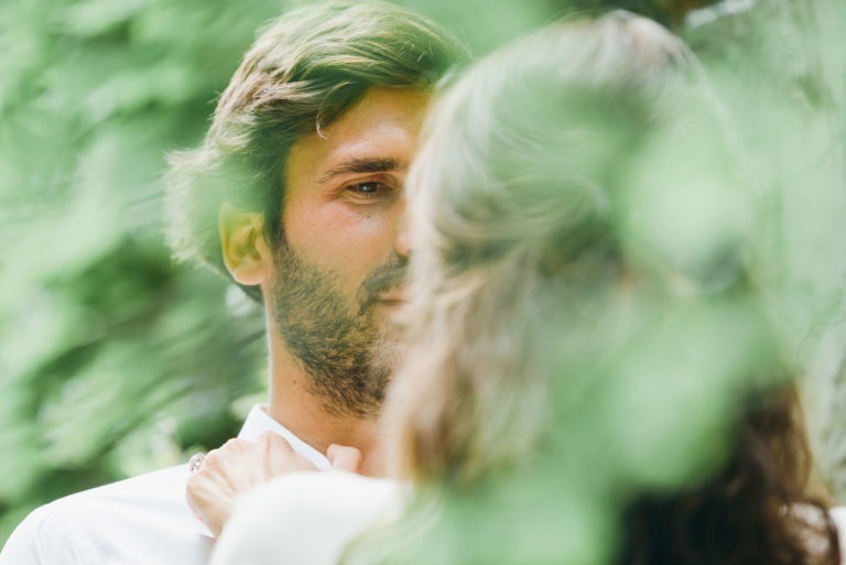 Gros plan d'un homme et d'une femme debout face à face, partiellement cachés par un feuillage vert. L'homme, aux cheveux châtain clair et à la barbe, regarde doucement la femme, qui tourne le dos à la caméra. Cette séance photo de couple dégage une atmosphère sereine et intime, avec une mise au point douce mettant en valeur la verdure et leur proximité.