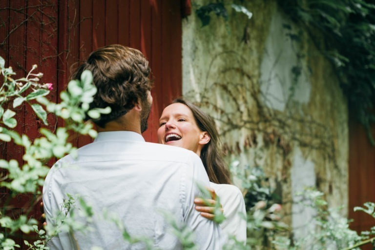 Une femme aux longs cheveux bruns et au sourire éclatant enlace un homme barbu aux cheveux bruns courts, tous deux vêtus de chemises blanches. Ils se tiennent devant un mur en bois rouge orné de lierre et d'autres feuillages verts, capturant un moment joyeux et intime dans cette séance photo de couple enchanteresse.