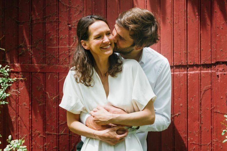 Une femme souriante en robe blanche se tient devant un mur en bois rouge patiné par les intempéries. Un homme en chemise blanche la serre dans ses bras par derrière et l'embrasse sur la joue. Tous deux semblent heureux et détendus, la lumière naturelle illuminant la scène, mettant en valeur la verdure à gauche - un moment parfait pour une séance photo de couple.