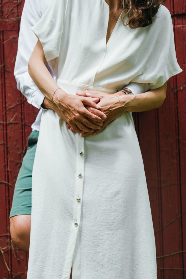 Une personne portant une robe blanche avec des boutons sur le devant se tient debout devant quelqu'un portant une chemise blanche et un short vert, ressemblant à un joli couple de séance de photos. La personne derrière a ses bras enroulés autour de celle de devant, lui tenant les mains. L'arrière-plan présente un mur en bois rouge avec une texture visible.