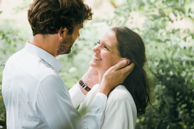 Un homme et une femme se tiennent côte à côte à l'extérieur, entourés d'une végétation luxuriante. L'homme touche doucement le visage de la femme avec ses deux mains et ils partagent un moment de joie, se souriant chaleureusement. Tous deux vêtus de chemises blanches, cette photo de séance de couple capture une scène sereine et intime.