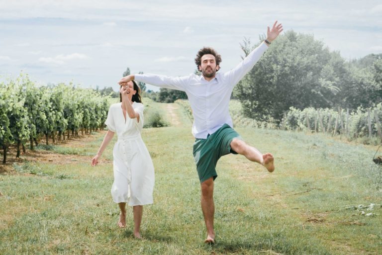 Un couple court joyeusement pieds nus dans un vignoble dans une belle séance photo de couple. L'homme, les bras écartés, affiche une expression enjouée en donnant un coup de pied en l'air. La femme, vêtue d'une robe blanche, sourit et lui tient la main. La verdure luxuriante du vignoble s'étend derrière eux sous un ciel partiellement nuageux.