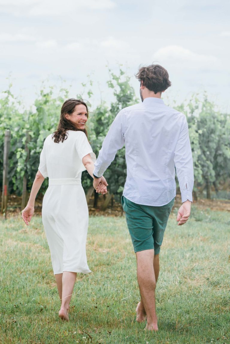 Un couple marche main dans la main dans un vignoble par une journée ensoleillée. La femme, en robe blanche, sourit à l'objectif tandis que l'homme, vêtu d'une chemise bleu clair et d'un short vert, regarde devant lui. Tous deux sont pieds nus, marchant sur l'herbe entre les rangées de vignes pendant leur séance photo de couple. Le ciel est clair et partiellement nuageux.