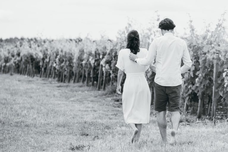 Une image en noir et blanc montre un couple s'éloignant à travers un vignoble. La femme porte une robe blanche et l'homme est habillé d'une chemise blanche et d'un short foncé. Ils sont bras dessus, bras dessous et marchent le long d'un chemin herbeux entre des rangées de vignes. La scène capture le romantisme paisible de leur séance photo de couple.