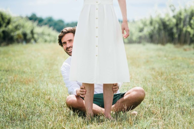 Un homme est assis en tailleur sur un terrain herbeux, souriant et tenant les chevilles d'une femme debout devant lui. La femme, vêtue d'une robe blanche boutonnée sur le devant, ajoute une touche d'élégance à cette scène décontractée et ludique. La verdure floue et la clôture en arrière-plan préparent le terrain pour leur séance photo de couple intime.