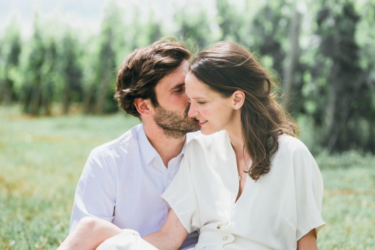 Un homme aux cheveux noirs et à la barbe embrasse doucement la joue d'une femme aux longs cheveux bruns. Ils sont assis côte à côte dehors sur l'herbe, entourés de verdure. La femme, vêtue d'une robe blanche, sourit doucement tandis que l'homme en chemise blanche termine leur séance photo romantique de couple.