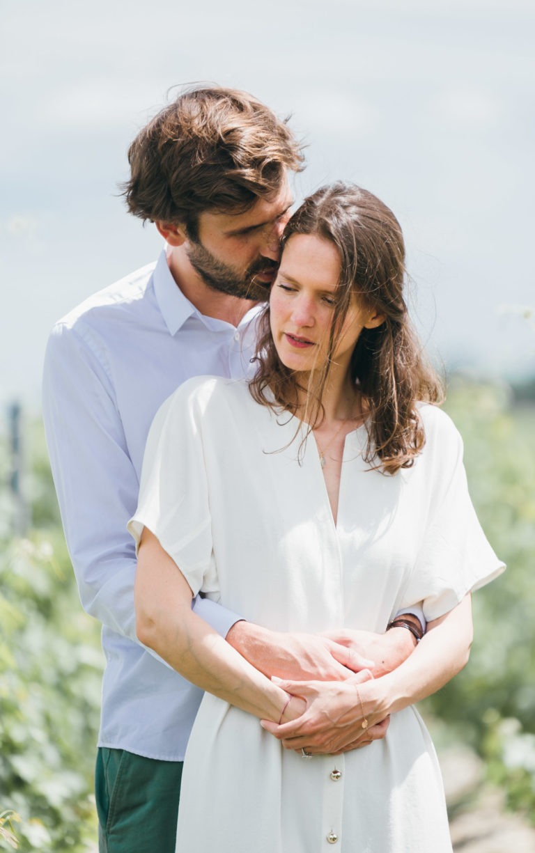 Un homme aux cheveux bruns et à la barbe enlace une femme aux longs cheveux bruns par derrière, sa joue reposant sur sa tête. La femme, vêtue d'une robe blanche, a les mains jointes sur les siennes. Ils se tiennent dans un décor extérieur avec de la verdure en arrière-plan sous un ciel nuageux, capturant une tendre séance photo de couple.