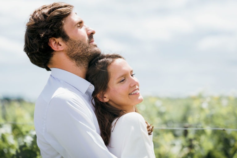 Un couple se tient dehors, s'embrassant et souriant. L'homme a les cheveux bruns courts et une barbe, et porte une chemise claire. La femme a les cheveux bruns longs et ondulés et un haut blanc, penchée en arrière vers l'homme. Ils se tiennent dans un champ ensoleillé avec un feuillage vert et un ciel bleu avec des nuages épars en arrière-plan - une véritable séance photo de couple.