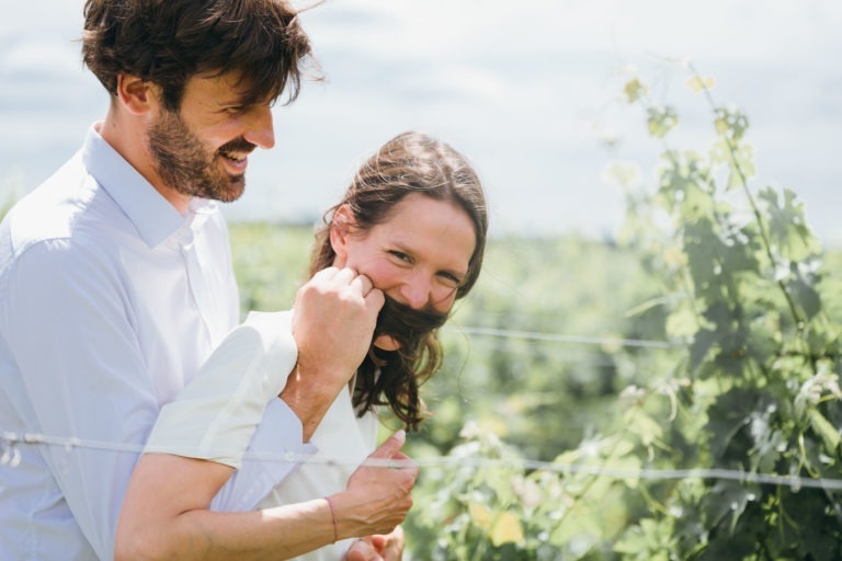 Un homme et une femme se tiennent ensemble dans un vignoble verdoyant et vibrant. L'homme, vêtu d'une chemise blanche, enlace la femme par derrière, tenant de manière ludique une mèche de ses cheveux sur son visage comme une moustache. Tous deux sourient et semblent heureux pendant leur séance photo de couple. L'arrière-plan est rempli de vignes et d'un ciel lumineux.