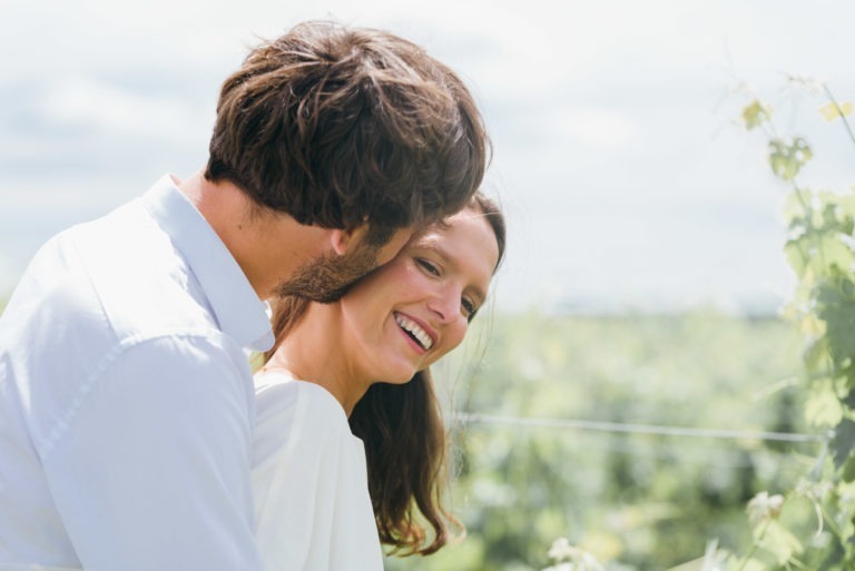 Un couple vêtu de blanc est dehors dans un vignoble verdoyant et luxuriant. L'homme, aux cheveux bruns et à la barbe, se penche pour murmurer à l'oreille de la femme aux longs cheveux bruns. Elle sourit joyeusement, regardant vers le bas et vers sa droite. Le soleil brille de mille feux, projetant une lumière chaleureuse sur cette belle séance photo de couple.