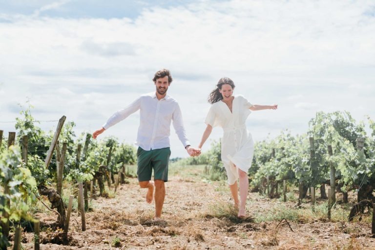 Un couple souriant traverse un vignoble ensoleillé en se tenant la main pendant leur séance photo de couple. L'homme porte une chemise blanche et un short vert, tandis que la femme est vêtue d'une robe blanche. Les rangées de vignes bordent leur chemin et le ciel est partiellement nuageux, créant une atmosphère joyeuse et insouciante.