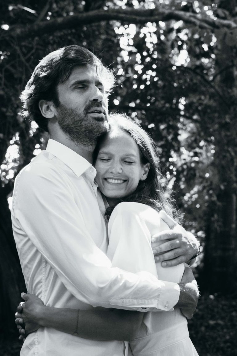 Une photo en noir et blanc montre un couple souriant qui s'embrasse en plein air. L'homme, à gauche, a les cheveux courts et une barbe, et porte une chemise à manches longues. La femme, à droite, a les cheveux longs et porte également une chemise à manches longues, posant sa tête sur la poitrine de l'homme. La lumière du soleil filtre à travers les arbres à l'arrière-plan de cette séance photo de couple.