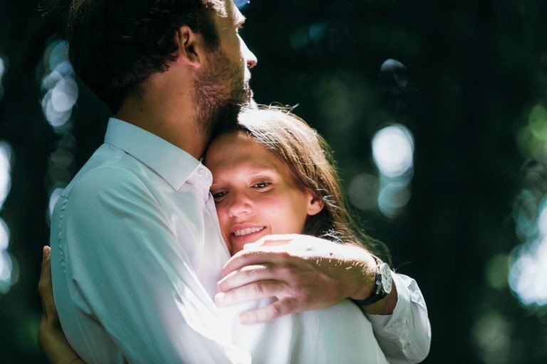 Un homme et une femme s'embrassent étroitement en plein air sur cette photo de couple lors d'une séance de spiritisme. L'homme aux cheveux courts et à la barbe regarde vers la gauche, portant une chemise blanche. La femme aux cheveux longs sourit avec contentement, les yeux fermés, et s'appuie contre son torse, son visage illuminé par la douce lumière du soleil filtrée à travers les arbres en arrière-plan.