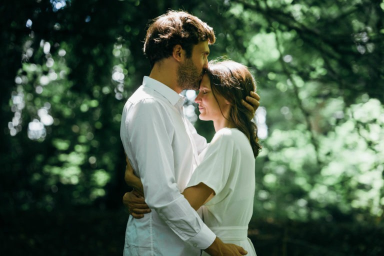 Un couple se tient près d'eux dans une forêt, capturé dans un moment intime. Tous deux vêtus de blanc, l'homme embrasse doucement le front de la femme tout en la prenant dans ses bras. La lumière du soleil filtre à travers les arbres, créant une atmosphère sereine et romantique autour d'eux, parfaite pour une séance photo de couple.