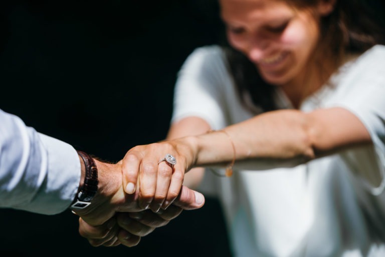 Une femme souriante portant un haut blanc est vue de près, tenant la main d'une personne portant une chemise blanche et une montre pendant leur séance photo de couple. Elle arbore une bague de fiançailles proéminente sur sa main gauche. L'arrière-plan est flou et sombre, attirant l'attention sur l'interaction et la bague.