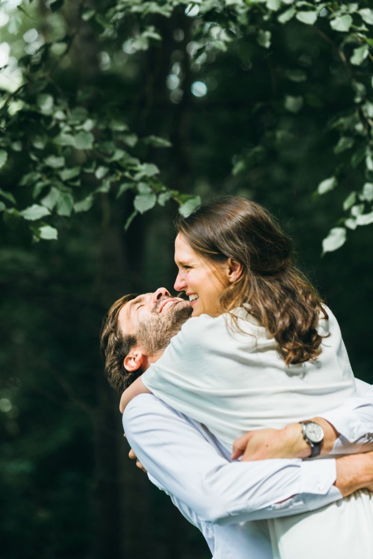 Un couple s'embrasse joyeusement dans un décor extérieur, entouré d'arbres et de feuillages verdoyants. L'homme, portant une chemise blanche et une montre-bracelet, soulève la femme, également vêtue de blanc, alors qu'ils partagent un rire pendant leur séance photo de couple. Les cheveux bruns de la femme tombent en cascade sur ses épaules.