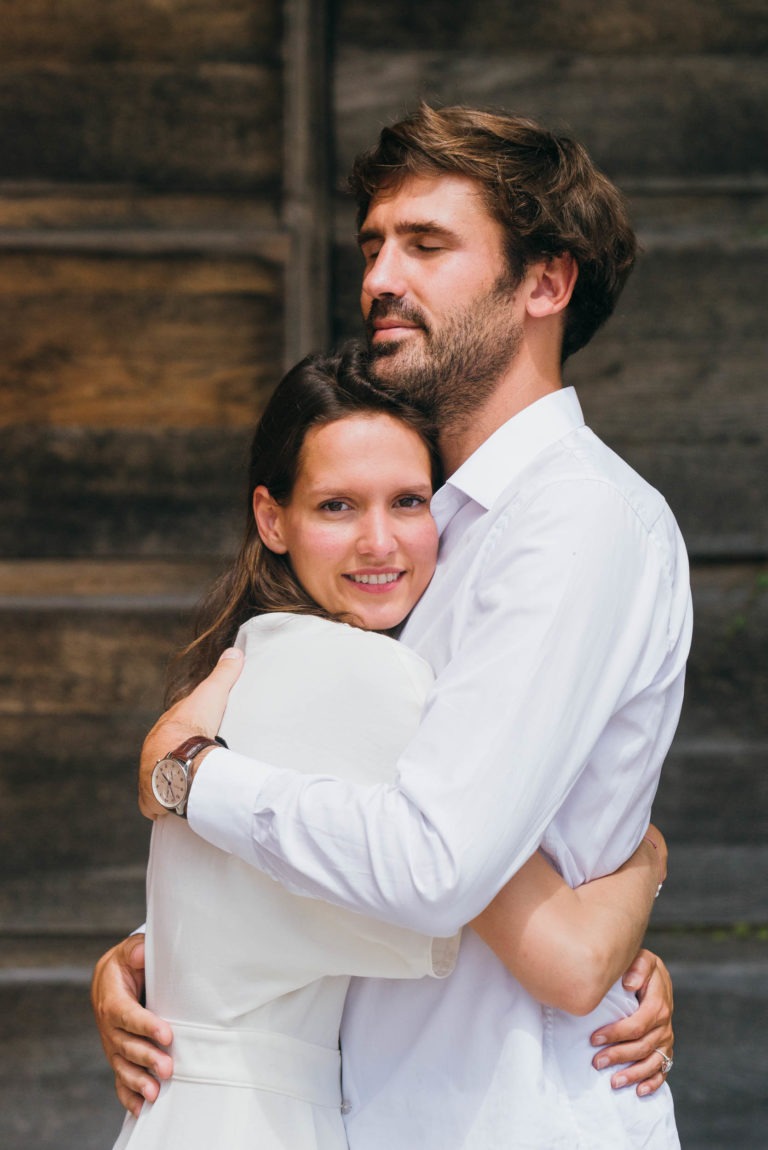 Un couple se tient debout à l'extérieur devant un décor en bois, l'un enlacé l'autre. La femme sourit doucement à la caméra tandis que l'homme, les yeux fermés, l'entoure de ses bras, semblant satisfait et paisible. Tous deux sont vêtus de chemises blanches ; c'est une séance photo de couple chaleureuse et intime.