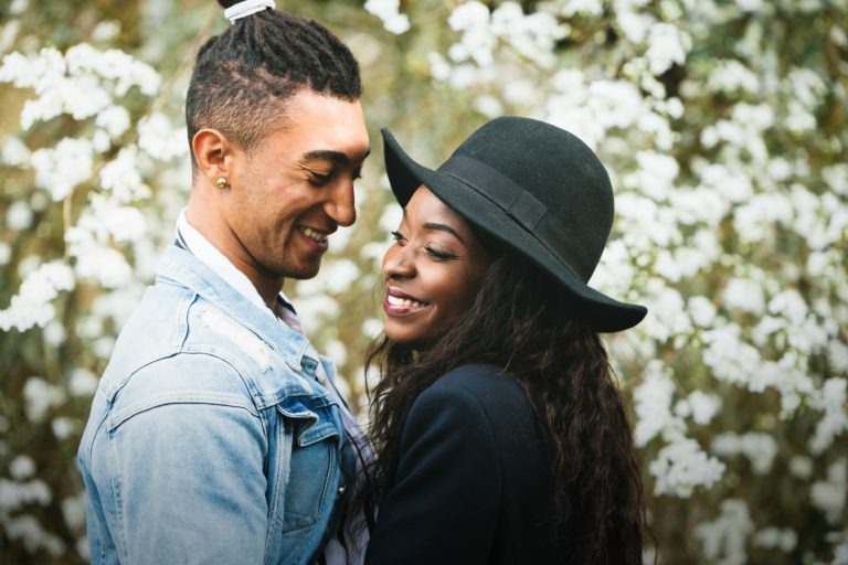 Un couple souriant se tient debout à proximité l'un de l'autre à l'extérieur. L'homme, vêtu d'une veste en jean bleu clair, et la femme, vêtue d'une veste sombre et d'un chapeau noir, partagent un moment d'affection. Des fleurs blanches en fleurs remplissent l'arrière-plan, créant une atmosphère romantique et sereine rappelant des séances intimes.