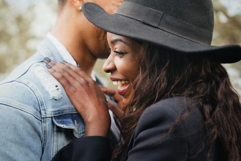 Une femme souriante, coiffée d'un chapeau et d'une veste noirs, se tient debout à côté d'une personne portant une veste en jean, la main posée sur son épaule. L'éclairage chaleureux et l'arrière-plan légèrement flou créent une atmosphère intime qui met en valeur l'expression joyeuse du couple, rappelant les moments partagés lors de séances de spiritisme.