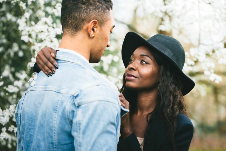 Une femme portant une veste sombre et un chapeau noir regarde avec amour un homme portant une veste en jean clair. Ils se tiennent l'un près de l'autre dans un jardin avec des fleurs blanches en arrière-plan, ressemblant à un moment intime souvent vu dans les séances de spiritisme. La femme a ses mains posées doucement sur les épaules de l'homme.