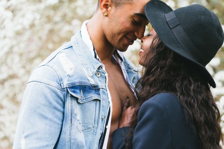 Un couple se blottit étroitement dans un décor naturel flou. L'homme porte une veste en jean et une chemise blanche, les boutons du haut défaits. La femme, aux cheveux longs et ondulés, porte un chapeau noir et un blazer. Ils se penchent, se touchent le nez et sourient, partageant un moment intime et affectueux qui rappelle les séances de spiritisme intemporelles.