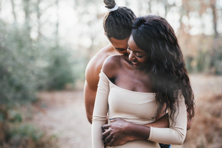 Un couple se tient dans une étreinte intime dans un décor extérieur boisé. La femme, souriante et vêtue d'une robe beige à épaules dénudées, est tenue par derrière par l'homme torse nu aux dreadlocks attachées. À proximité, des vestiges de séances passées persistent au milieu de la verdure floue de la forêt et d'un sentier couvert de feuilles mortes.