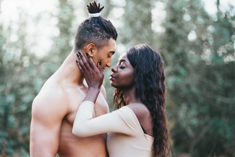 Un homme torse nu avec un chignon et une femme portant un haut beige à épaules dénudées se tiennent près l'un de l'autre dans une étreinte, se regardant dans les yeux. La femme pose doucement sa main sur le visage de l'homme, comme dans une séance intime de connexion, en plein air avec de la verdure floue en arrière-plan.