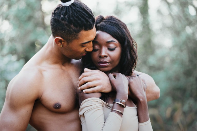 Un homme torse nu aux cheveux tressés enlace une femme par derrière, posant son menton sur son épaule. La femme, vêtue d'un haut beige à épaules dénudées et à manches longues, regarde vers le bas avec une expression sereine. Ils se tiennent dehors devant un arrière-plan de feuillage vert flou, transmettant un sentiment d'intimité rappelant les séances de spiritisme de couple.