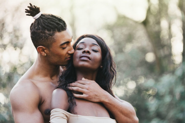 Un homme torse nu avec des dreadlocks embrasse la joue d'une femme aux cheveux longs et ondulés tout en la prenant dans ses bras. La femme ferme les yeux et incline la tête en arrière, posant sa main sur son épaule. En extérieur, sur un fond de nature floue et verdoyante, la scène respire l'intimité sereine souvent observée lors des séances de couples amoureux.