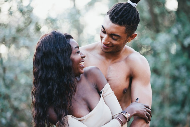 Une femme souriante aux cheveux longs et ondulés, portant un haut à épaules dénudées, serre dans ses bras un homme torse nu aux cheveux tressés et coiffé d'un élastique blanc. L'homme la regarde affectueusement. Ils se tiennent dehors, avec une végétation floue en arrière-plan, rappelant une scène de séances de spiritisme romantiques que les couples chérissent pour toujours.