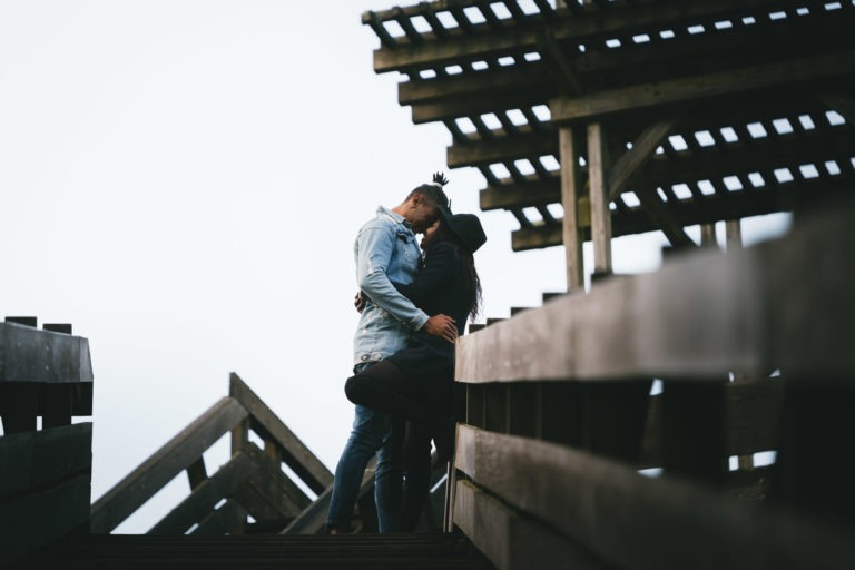 Un couple s'embrasse passionnément sur un pont en bois surplombé par une structure en treillis. L'homme, vêtu d'une veste en jean légère, se penche pour le baiser, tandis que la femme, coiffée d'un chapeau, l'entoure. Le ciel brumeux et couvert renforce l'atmosphère intime et sereine souvent ressentie lors des séances privées entre couples.