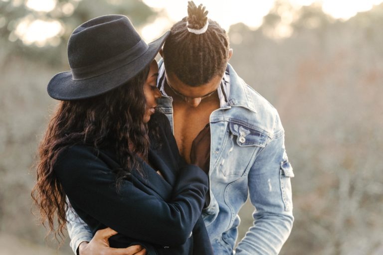 Un couple s'embrasse en plein air. La femme, qui porte un chapeau et un manteau noirs, regarde affectueusement l'homme dans les yeux. L'homme, les cheveux tressés noués en chignon et portant une veste en jean légère, la regarde. Leur moment intime et tendre semble presque éthéré, comme s'il faisait partie d'une séance de spiritisme intemporelle.