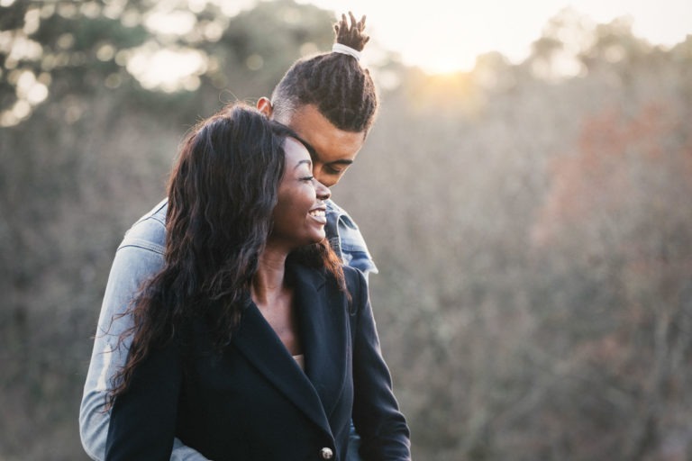 Un couple se tient dehors, devant un décor naturel composé d'arbres. La femme, aux cheveux longs et ondulés, sourit tout en portant un manteau sombre. L'homme, aux dreadlocks courtes attachées en queue de cheval, l'embrasse doucement par derrière, sa tête reposant près de la sienne. Ce moment de tendresse pourrait être le point culminant de nombreuses séances pour les couples en quête de connexion.