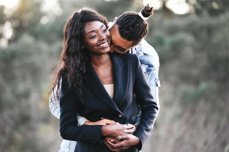 Un couple s'embrasse en plein air. La femme, vêtue d'un blazer et d'une chemise sombres, sourit tandis que l'homme, vêtu d'une veste en jean et coiffé d'un chignon, la serre dans ses bras par derrière et se penche vers lui. Au milieu de la verdure floue de leur environnement naturel, ils dégagent une connexion harmonieuse souvent observée lors de séances intimes de couples.