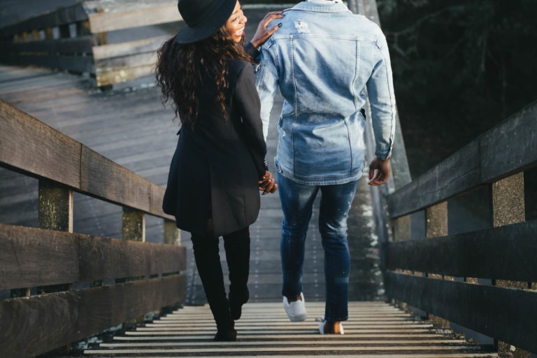 Un couple descend des escaliers en bois sur un pont. La personne à gauche a les cheveux longs, porte un chapeau noir, un manteau noir et un pantalon noir. La personne à droite porte une veste en jean bleu clair, un jean et des chaussures blanches. Se tenant la main et se souriant, ils semblent avoir partagé des moments privés proches de séances intimes.