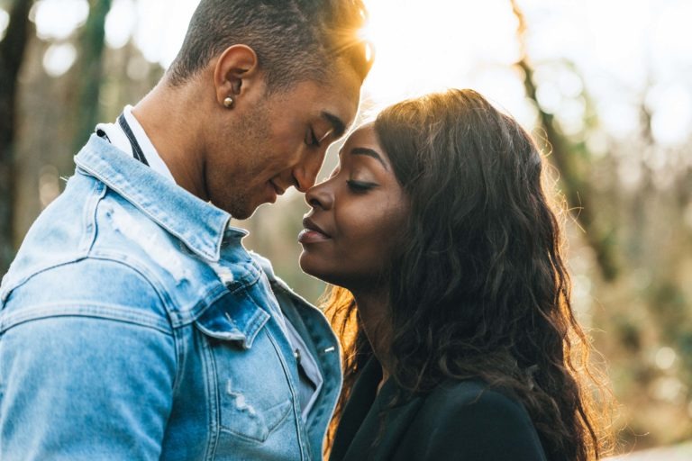 Un couple se tient debout, le front contre le mur, les yeux fermés, dans un moment d'intimité. L'homme, vêtu d'une veste en jean bleu clair, a un léger sourire. La femme, vêtue d'une tenue sombre, a de longs cheveux bouclés qui tombent sur ses épaules. C'est comme si leur connexion était presque spirituelle alors que le soleil brille doucement à travers les arbres en arrière-plan.