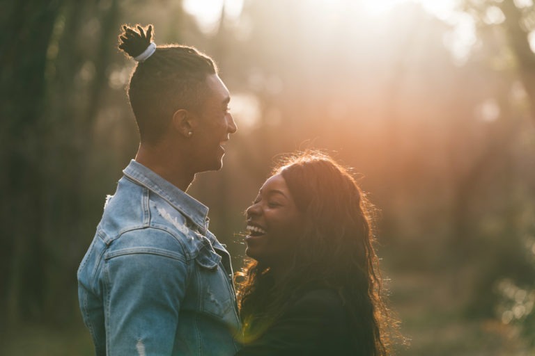 Un couple se tient face à face dans une forêt ensoleillée. L'homme, coiffé d'un chignon et vêtu d'une veste en jean, sourit à la femme aux cheveux longs et ondulés qui rit. La lumière du soleil filtre à travers les arbres, créant une lueur chaude et dorée autour d'eux, un moment paisible qui fait écho à l'intimité des séances de spiritisme avec ses proches.