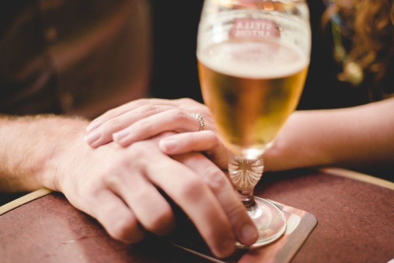 Deux mains se tiennent doucement sur une table en bois du Café Brun Bordeaux, avec un verre de bière sur un dessous de verre au premier plan. Les mains semblent appartenir à un couple, l'un portant une bague en argent. L'accent est mis sur le geste intime, avec le verre de bière légèrement flou à l'arrière-plan.